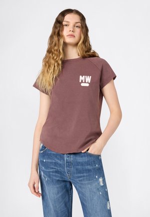 Jeune femme aux longs cheveux ondulés portant un t-shirt mauve à manches courtes avec le logo « MW » et un jean bleu déchiré, main dans la poche, fond neutre.