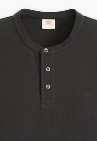 Crna Henley majica od pamuka. Ima okrugli izrez, dugmad na prednjoj strani s pet dugmadi i diskretni logo izvezan na prsima.