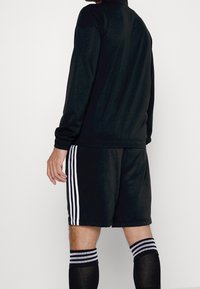 Schwarzer sportlicher Jogginganzug bestehend aus einem langärmeligen Oberteil und Shorts mit drei weißen Streifen an den Seiten, kombiniert mit gestreiften knielangen Socken.