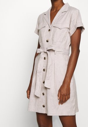 Krótka sukienka beżowa z krótkim rękawem, zapinana na guziki, z przednimi kieszeniami i paskiem w talii, noszona przez osobę o ciemnej karnacji na jednolitym tle.
