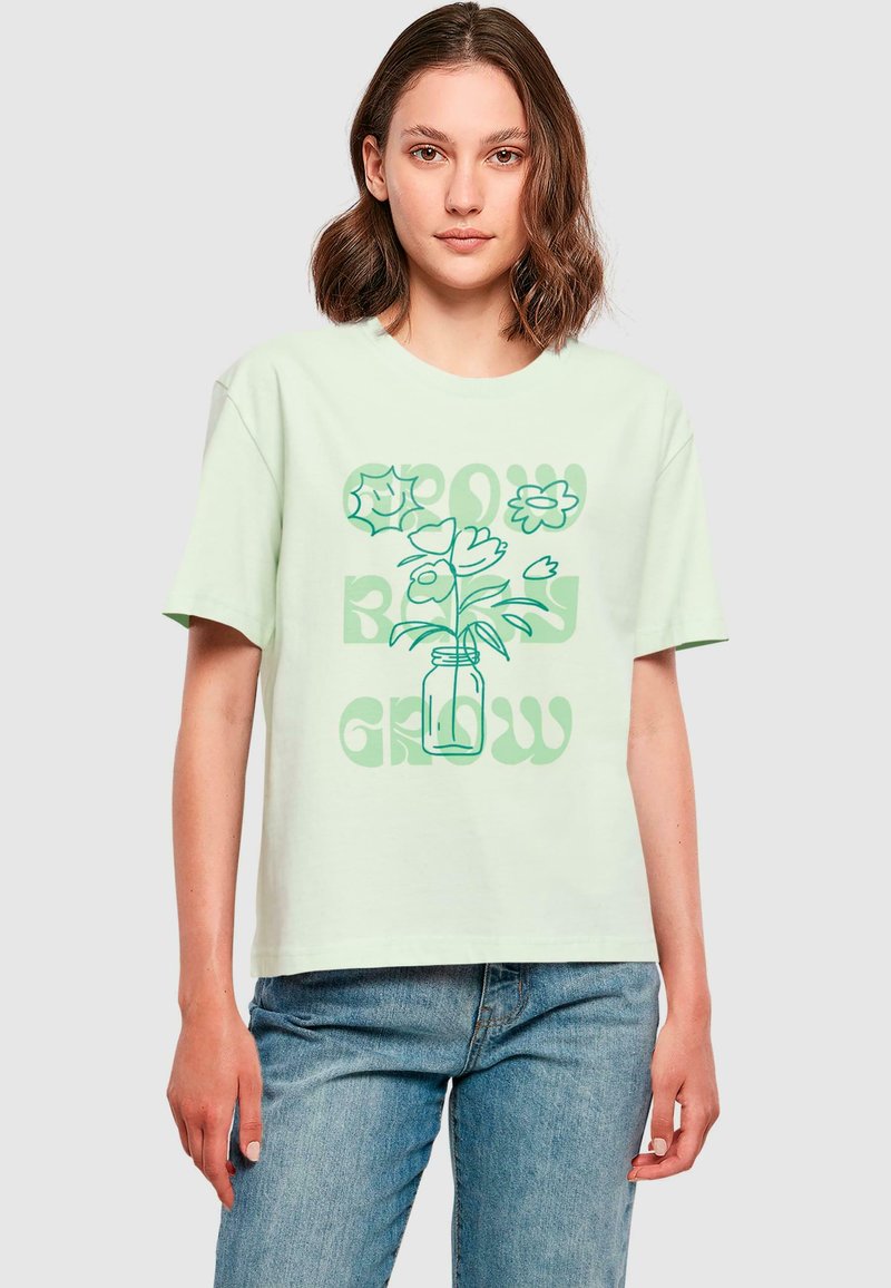 Hellgrünes Baumwoll-T-Shirt mit kurzen Ärmeln, das ein Grafiken von Blumen in einem Glas und den Text "WACHSTUM" in stilisierter Schrift aufweist.