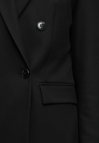 Μέρος μαύρου προσαρμοσμένου σακακιού που δείχνει δύο μαύρα κουμπιά και μια τσεπάκι με καπάκι σε λεία ύφασμα.