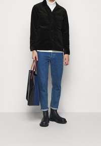 Giacca in velluto a coste nera con due tasche sul petto, dolcevita bianco, jeans blu, stivali Chelsea neri e una borsa a tracolla multicolore.