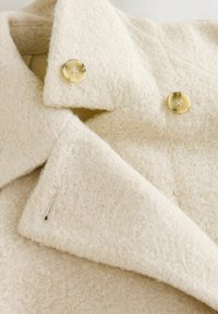 Krémovo sfarbený textured kabát z mäkkého, chlpatého materiálu, s širokým golierom a dvoma žltými gombíkmi vpredu.