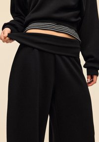 Sort loungewear-sæt af stof med en løstsiddende top og bukser med vide ben. Toppen har et lagdelt udseende med en gråstribet bånd ved taljen.