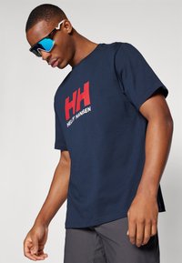 Navy plava pamučna majica s odvažnim crvenim logotipom "HH" i tekstom "HELLY HANSEN" sprijeda. Model nosi bijele i plave sunčane naočale.