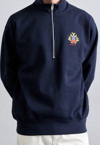 Person wearing a navy blue quarter-zip sweatshirt with a multicolored Bally crest embroidered on the chest, paired with light pants.
