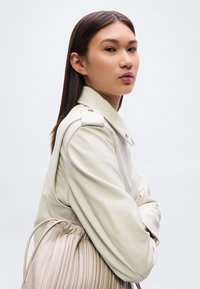 Δερμάτινο μπουφάν ανοιχτό μπεζ με επιθέματα στους ώμους, με λεία υφή. Το μοντέλο φορά μια πλισέ τσάντα ταφ με κορδόνι που ταιριάζει με το ντύσιμο.