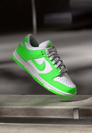 Baskets Nike blanches et vert néon avec lacets gris suspendues au-dessus d'une surface en béton sur un fond sombre flou.