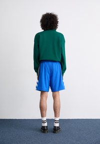 Personne aux cheveux bouclés portant un pull vert, un short de sport bleu, des chaussettes blanches à rayures noires et des chaussures noires, debout contre un mur blanc.