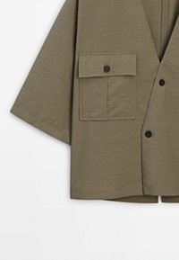 Chemise vert olive à manches courtes avec une grande poche boutonnée à l'avant et des boutons noirs sur le devant.