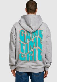 Szürke kapucnis pulóver, melyen merész, színes "GAME TIME GRIT" felirat található türkiz színben, narancssárga körvonalakkal. Pamut anyagból készült, laza szabású.