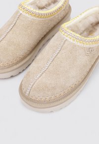 Une paire de pantoufles en suédine beige à enfiler, ornées de bordures brodées jaunes et blanches, avec une doublure en fourrure douce et des semelles en caoutchouc.