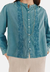 Helles blaues Jeanshemd mit Stehkragen, verziert mit aufwendiger floralem Stickerei entlang der Vorderseite und Knopfverschluss, kombiniert mit beigen gestreiften Hosen.