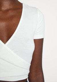Gros plan d'une personne portant un crop top blanc à manches courtes portefeuille avec un profond décolleté en V.