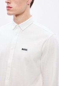 Biela košeľa s golierom z textúrovaného materiálu. Na ľavej strane hrudníka je čierne vyšívané logo "BOSS". Čistý, zapínací dizajn.