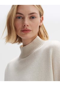 Cremfarbener Strickpullover mit hohem geripptem Kragen, der eine weiche Textur und ein dezentes Muster im Strickdesign aufweist.