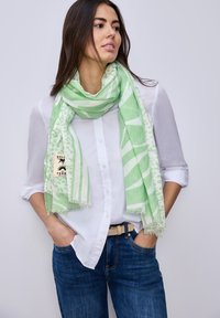 Femme portant une chemise blanche boutonn ée et un jean bleu, les mains dans les poches, enveloppée d'une écharpe légère verte à motifs.