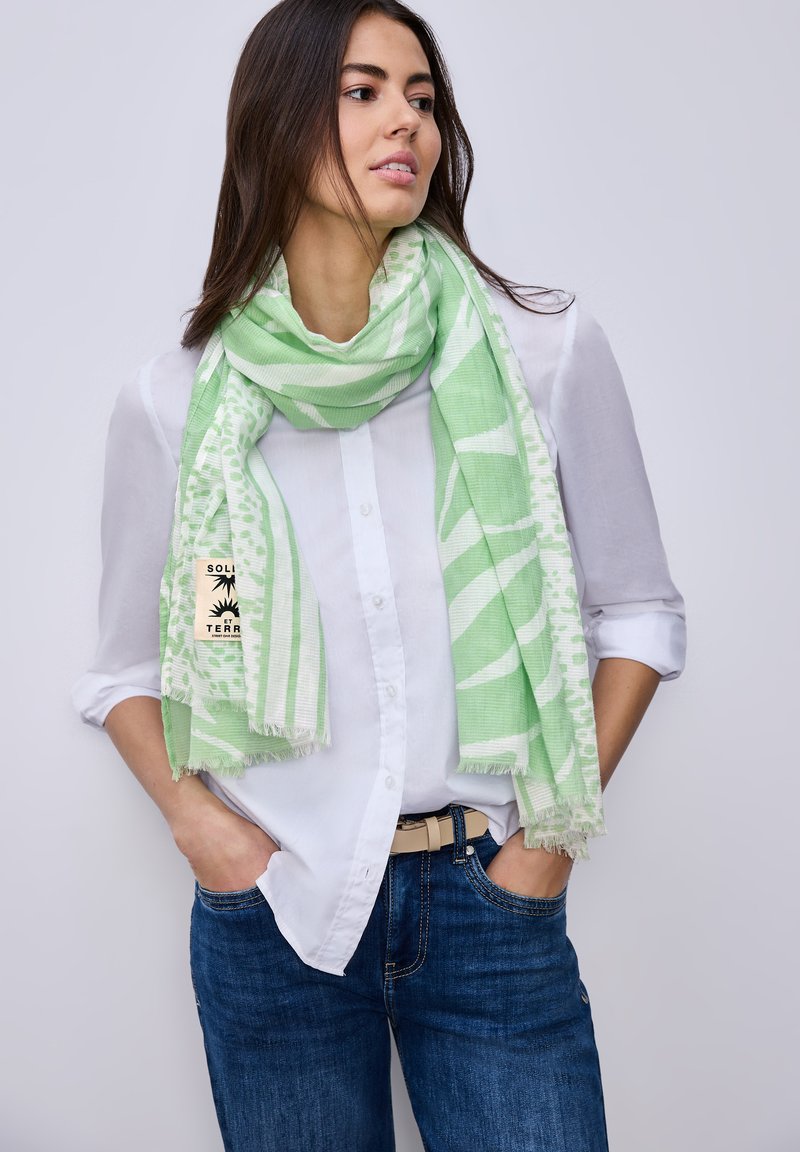 Femme portant une chemise blanche boutonnée et un jean bleu, les mains dans les poches, enveloppée d'une écharpe légère verte à motifs.