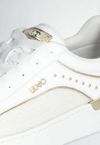 Sneaker bianca con tessuto testurizzato, accenti dorati e un logo in metallo sulla linguetta. Presenta lacci piatti e piccoli chiodi dorati lungo il lato.