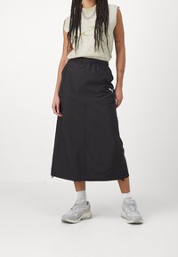 Mustat keskikokoiset hameet, joissa on joustava vyötärö ja vetoketjut helmassa, yhdistettynä vaaleanvihreään hihattomaan toppiin ja harmaisiin tennareihin.