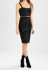 Černý dvoudílný kostým složený z krátkého topu a přiléhavé tužkové sukně, zdobený detaily ve tvaru nýtu vytvářejícími geometrické vzory.