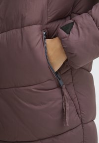 Puffjacka i mörk mauve med en dragkedjeficka. Har en kviltad yta och en triangulär svart logopatch på ärmen.
