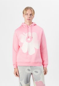 Rosa Kapuzenpullover mit einem großen weißen Blumenmotiv. Weiches Material, lange Ärmel und Kapuze mit Kordelzug. Kombiniert mit gemusterten grauen Hosen.