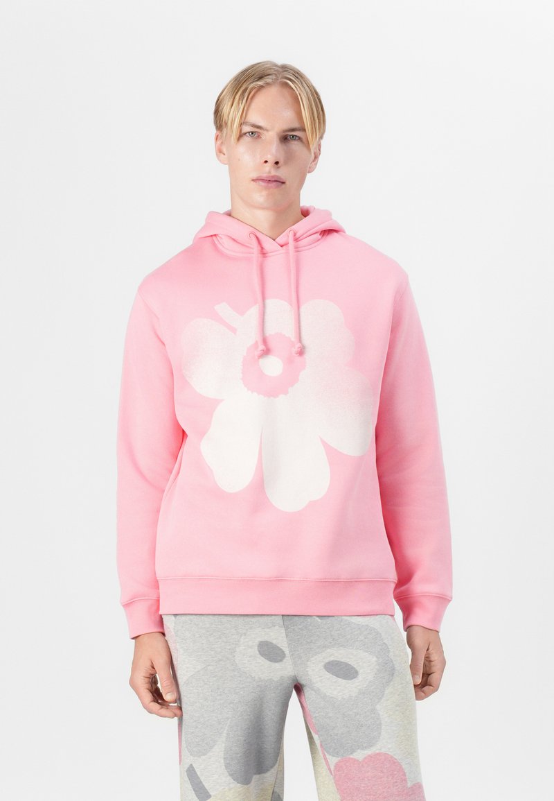 Rosa Kapuzenpullover mit einem großen weißen Blumenmotiv. Weiches Material, lange Ärmel und Kapuze mit Kordelzug. Kombiniert mit gemusterten grauen Hosen.