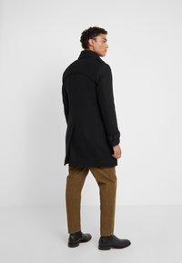 Schwarzer Wollmantel mit hohem Kragen, tailliertem Design und geknöpften Manschetten. Kombiniert mit braunen Cordhosen und schwarzen Stiefeletten.
