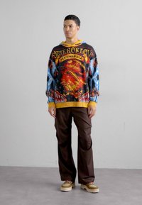 Multicolored knit hoodie with intricate patterns and a yellow hem, paired with brown wide-leg trousers and beige platform shoes.