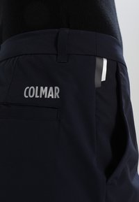 Gros plan sur un pantalon noir avec une boucle de ceinture, une poche arrière et le logo "COLMAR" imprimé en blanc sur le côté supérieur gauche.
