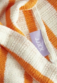 Pletiva s izmjeničnim narančastim i bijelim prugama, s teksturiranim uzorkom i malom ljubičastom etiketom na kojoj piše "JAXX."