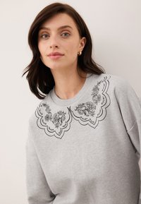 Kobieta z średniej długości brązowymi włosami, ubrana w szary sweter z czarnym haftem kwiatowym na kołnierzu i małymi kolczykami w kształcie kółka.