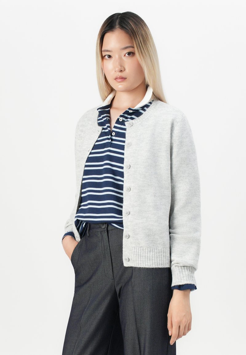 Cardigan a maglia grigio chiaro con collo rotondo, chiusura a bottoni e polsini a coste, indossato sopra una polo a righe blu navy, abbinato a pantaloni scuri.