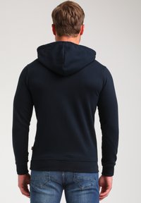 Sudadera azul marino con capucha ajustable, mangas largas y puños acanalados. Tejido suave con una textura lisa y una etiqueta en la parte inferior izquierda.
