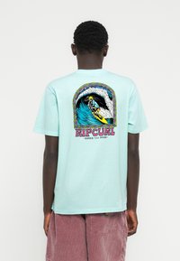 Μια βαμβακερή μπλούζα σε μέντα πράσινο χρώμα έχει ένα πολύχρωμο γραφικό με έναν σέρφερ πάνω σε κύμα, με το κείμενο «RIPCURL - SHRED TILL DEAD» από κάτω.