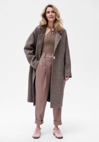 Bruine jas met een visgraatpatroon, beige ribbeltop, taupe broeken met opgerolde manchetten en lichtroze sneakers. Gladde texturen.