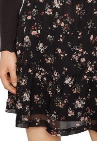 La mano di una donna accanto a un vestito floreale nero con fiori rosa e bianchi, caratterizzato da un orlo arricciato e dettagli in tessuto trasparente vicino al ginocchio.