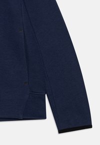 Námořnická modrá mikina s texturovanou tkaninou, bočními rozparky, kontrastními černými akcenty na lemu a viditelnými švy.