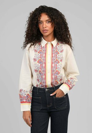 Femme aux cheveux bouclés portant un chemisier crème à motifs floraux rouges et violets, rentré dans un jean foncé taille haute.