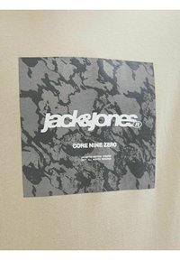 Beżowy t-shirt z czarnym graficznym kwadratem o teksturowanym wzorze i białym napisem "jack&jones CORE NINE ZERO" na środku.
