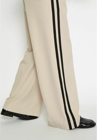 Beige wide-leg pants featuring black side stripes. Made of smooth fabric, they have pockets and are paired with shiny black footwear.
