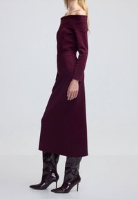 Robe longue bordeaux à épaules dénudées avec manches longues, taille cintrée et jupe fluide ; associée à des bottes à talons aiguilles noires brillantes.