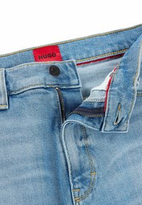 Ljusblå denimjeans med röd "HUGO"-etikett, dragkedjestängning, metallhardware, kontrasterande gul sömnad och en mjuk, texturerad yta.