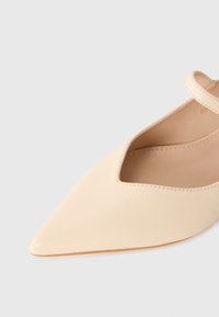 Beige pointed-toe flat shoe with a V-cut front and ankle strap, displayed on a light gray background.