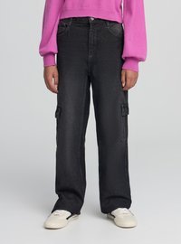 Osoba ubrana w czarne jeansy typu cargo, białe trampki i różowy sweter z długim rękawem, stojąca na tle jednolitego jasnego tła.