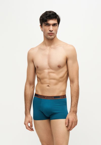 Jeune homme aux cheveux foncés portant un boxer bleu sarcelle avec une ceinture de marque Emporio Armani, debout devant un fond clair uni.