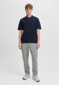 Giovane in piedi, indossa una polo a maniche corte blu scuro, pantaloni grigi chiari e sneakers argentate su uno sfondo bianco uniforme.