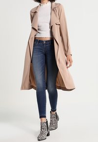 Abrigo trench beige con solapas anchas, combinado con una blusa blanca ajustada, jeans oscuros ajustados y botas de tobillo con estampado animal en blanco y negro.